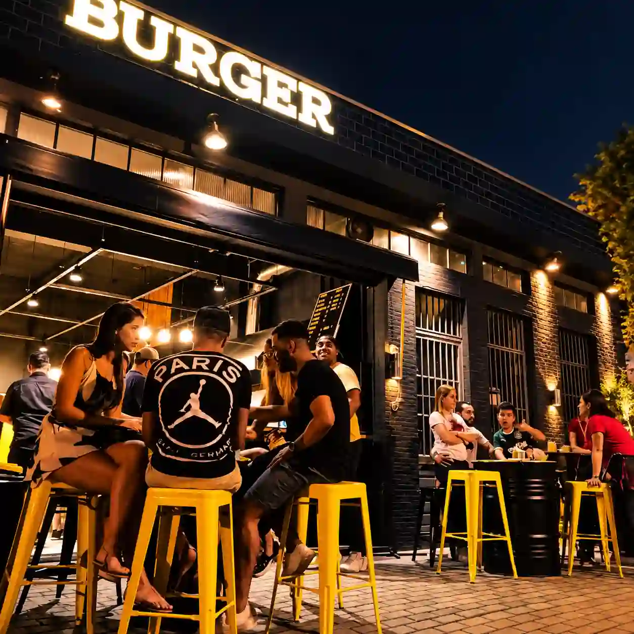 Área externa do The Vila Burger à noite, com banquetas amarelas e clientes reunidos